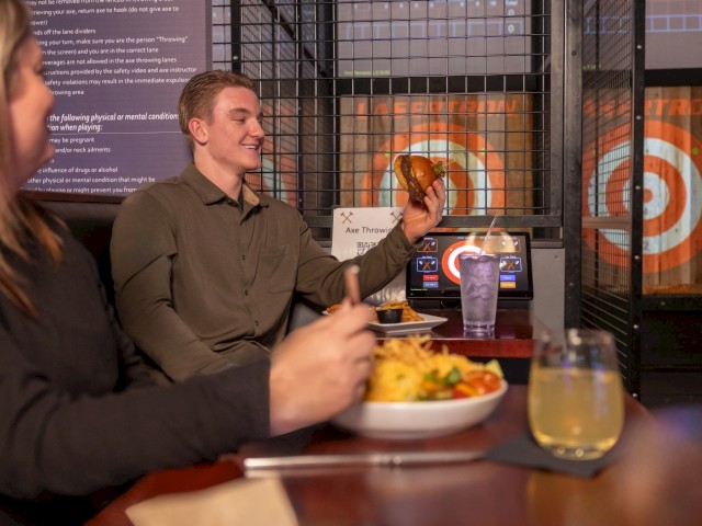 People enjoying a meal at a restaurant with axe throwing in the background.