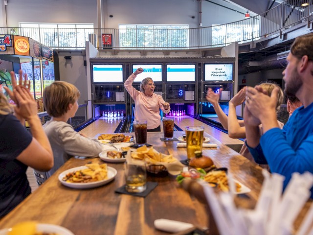 A group celebrates at a birthday party in a bowling alley with snacks, drinks, and a woman dancing or cheering in front of the pins.