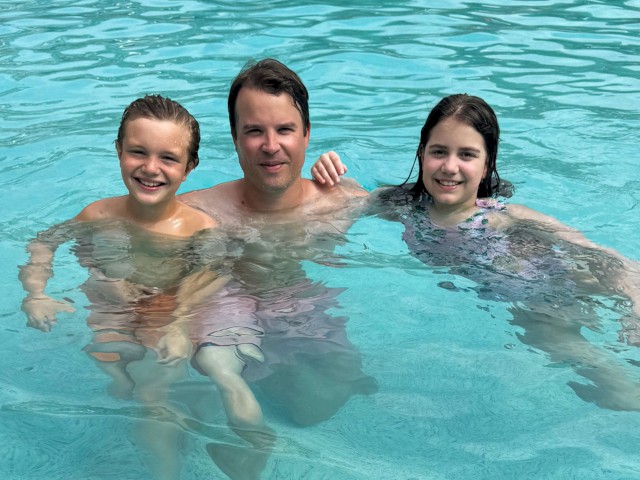 A man with two children swim and smile in a bright blue pool on a sunny day, enjoying a fun moment together.