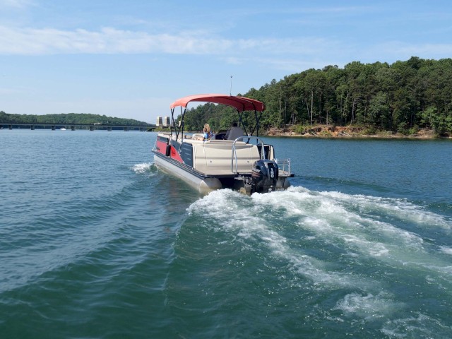 A small boat with a red canopy is moving across a calm, blue lake with a green, forested shoreline and a bridge in the distance.