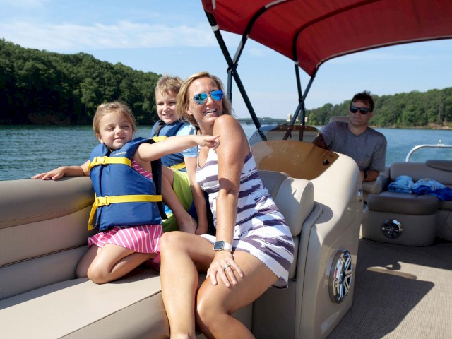 A woman and three children enjoy a sunny day on a boat, with the woman smiling and pointing, surrounded by water and green trees.