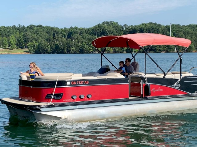 A red and black pontoon boat with people enjoying a lake day, surrounded by green trees and calm water under a partly cloudy sky.