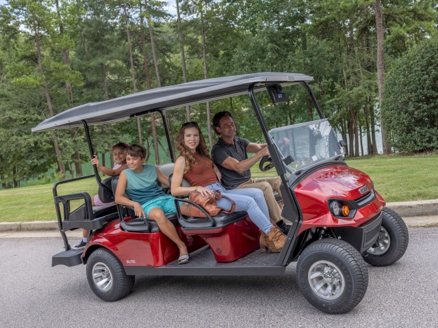 A family of four riding in a red golf cart on a gray paved road surrounded by lush green trees and grass.