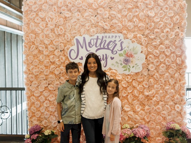 A woman with two children stands in front of a pink flower wall with a 