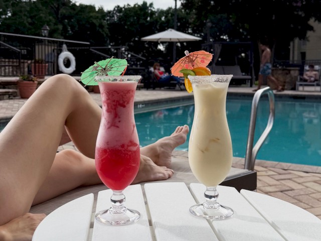 Two colorful tropical drinks with umbrellas are on a poolside table, while someone relaxes by the pool in the background.