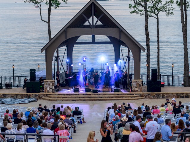 A band performs on an outdoor stage by a lake during sunset, with an audience seated in front, surrounded by trees and scenic water views.