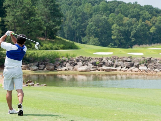 A golfer takes a shot on a lush green course near a water hazard with trees and sand traps in the background.