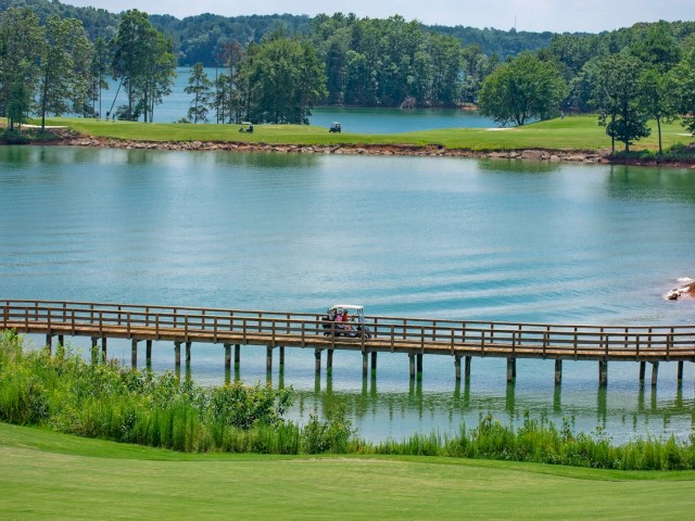 A golf course with a lake, a bridge, and a golf cart driving across it, surrounded by lush greenery and trees.