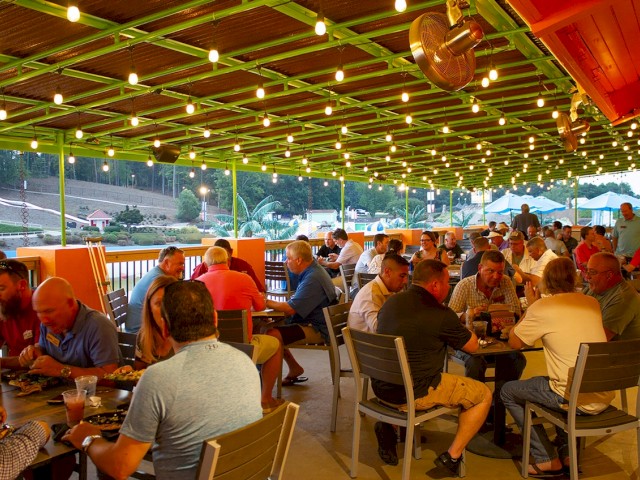 People are dining at an outdoor restaurant with bright string lights and a view of trees and hills in the background.
