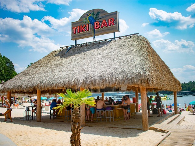 A tropical-style Tiki Bar on a sandy beach with people relaxing and enjoying drinks under a thatched roof.