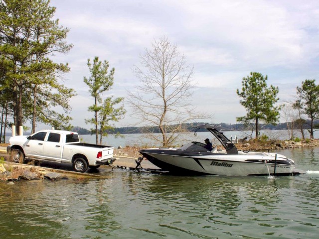 A pickup truck is towing a Malibu boat into a lake, with trees and cloudy sky in the background.