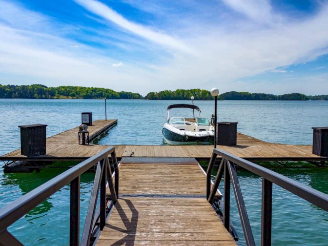 A wooden dock extends into a calm lake with a boat tied up, surrounded by trees under a partly cloudy sky.