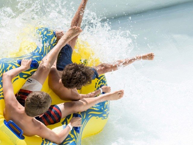 Three kids are having fun on a yellow inflatable raft, splashing water as they slide down a water slide.