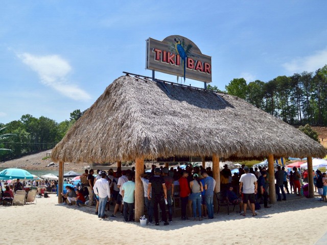 A beach scene with people gathered under a thatched-roof structure with a sign that reads 