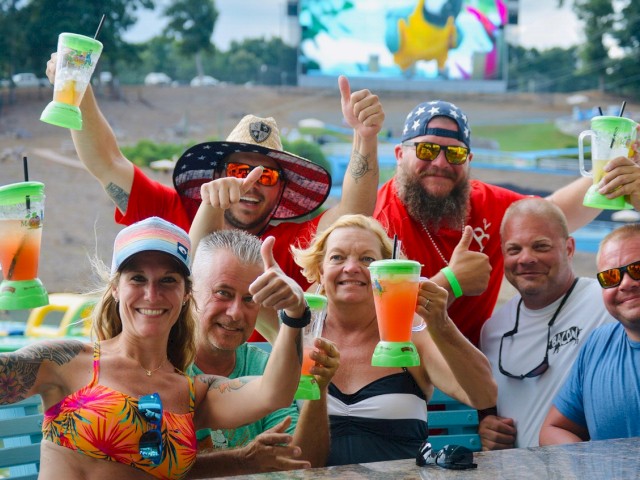 People celebrating with colorful drinks, outdoors in a group with thumbs up and smiles, with a large screen in the background showing an image.