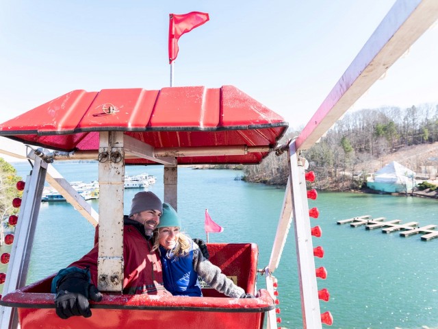 A couple is sitting in a red Ferris wheel cabin by a lake with docks in the background. They are smiling and appear to be enjoying it.
