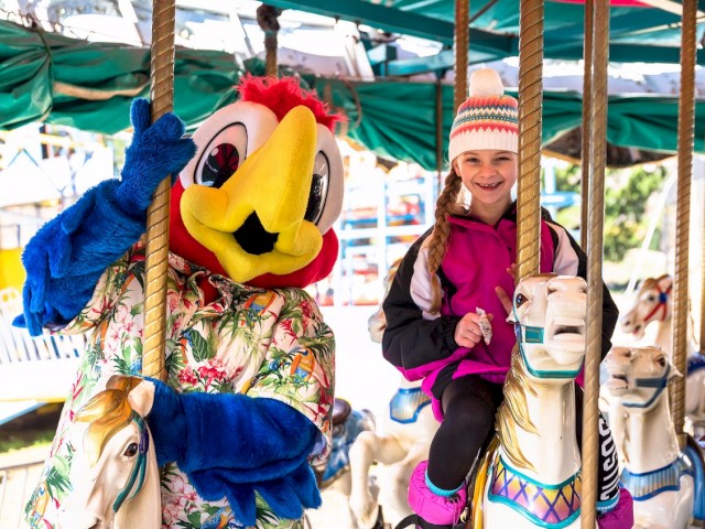 A person in a colorful bird costume and a smiling child are riding a carousel horse, enjoying a day at the amusement park.