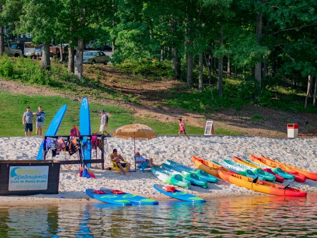 People relax and prepare for kayaking at a lakeside beach with colorful kayaks, paddleboards, and a shaded grassy area.