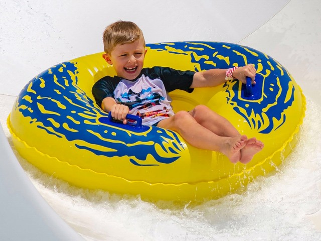 A boy happily rides a yellow and blue inflatable tube down a water slide.