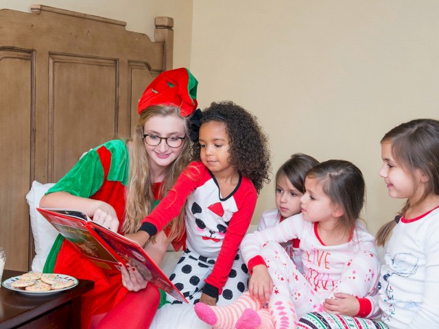 A person in a festive outfit reads a book to four children on a bed, with cookies and milk nearby.