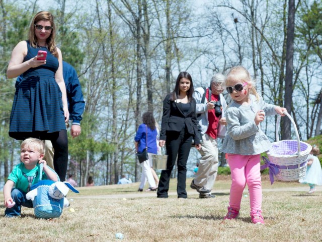 Children are participating in an outdoor Easter egg hunt while adults, including one on a smartphone, watch them.