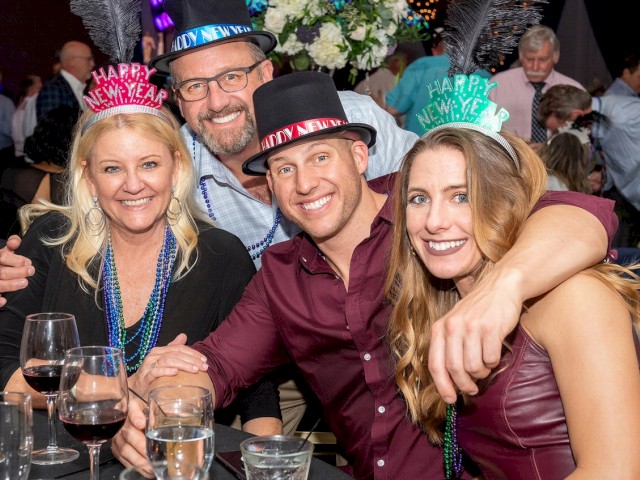 A group of four people wearing Happy New Year hats, smiling and celebrating at a party with drinks and decorations around them.