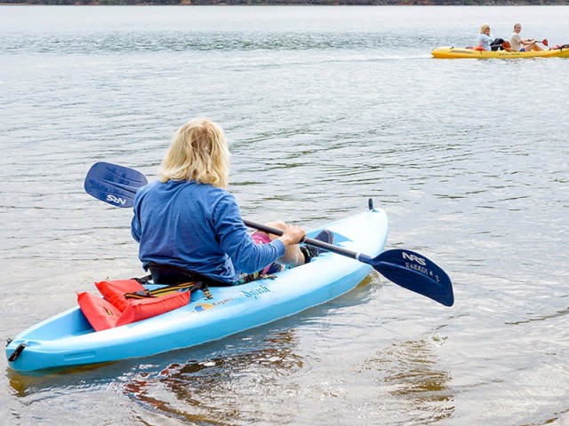A woman in a blue jacket paddles a blue kayak on a lake, with two people in a yellow kayak in the background enjoying a water activity.