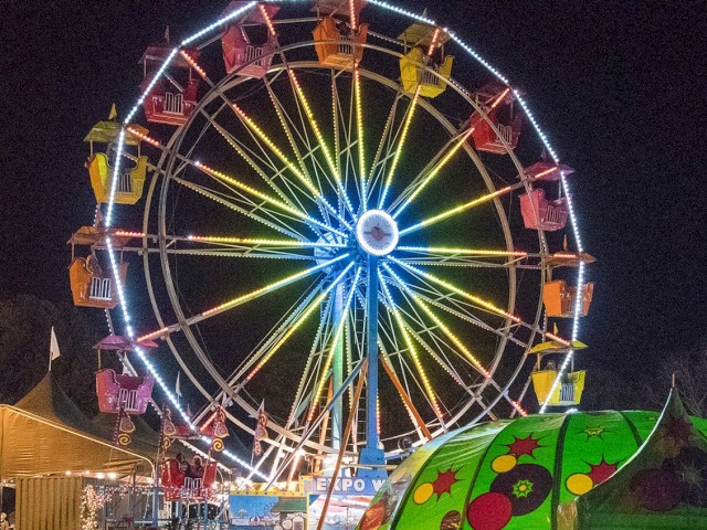 A colorful Ferris wheel lit up at night, with small amusement rides and tents visible below at a lively fair.