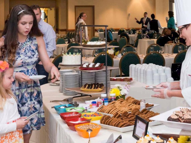 A buffet table with cookies, cakes, and toppings at a banquet; a woman and two young girls serve themselves while other guests chat nearby.