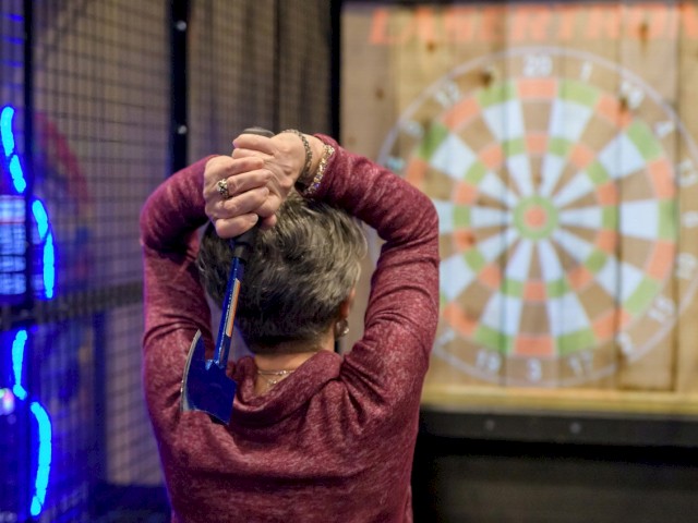 A person with gray hair wearing a red hoodie is about to throw a dart at a dartboard in an arcade or gaming area.