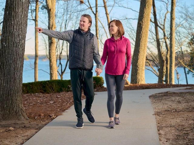 A man and woman are walking hand-in-hand on a paved trail through a wooded area near a lake, enjoying a sunny outdoor walk.