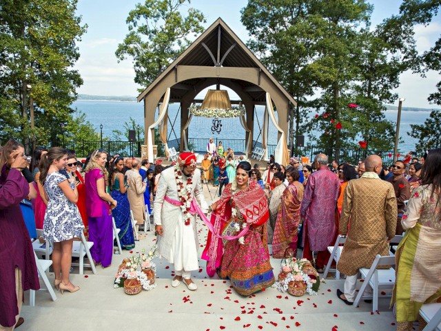 A couple in traditional attire is exiting an outdoor wedding ceremony, surrounded by seated guests, under a decorated structure near a body of water.
