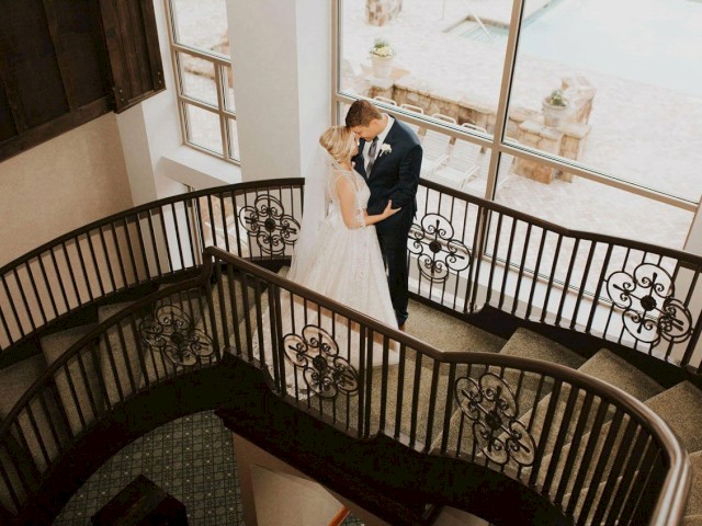 A couple in wedding attire shares a romantic moment on a beautifully wrought spiral staircase, with large windows in the background.