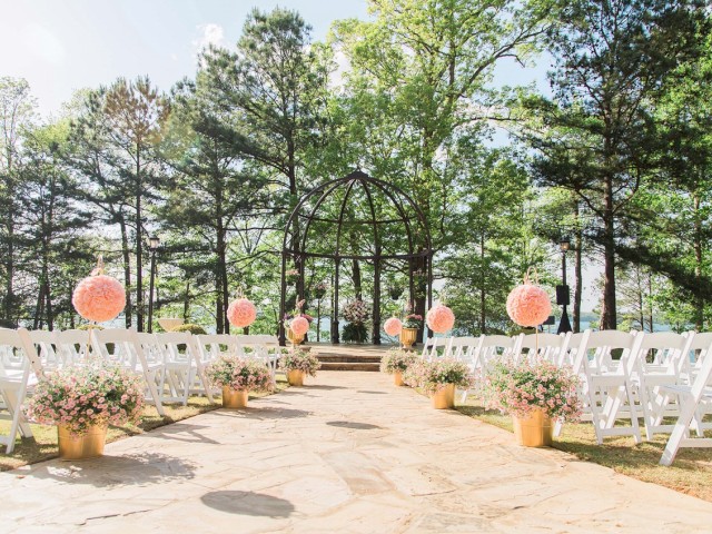 An outdoor wedding setup with white chairs, floral arrangements in gold vases lining the aisle, and a gazebo in the background.