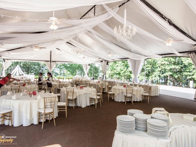 An elegantly decorated outdoor event tent with round tables covered in white linens, chandeliers, and place settings, set up for a formal gathering.