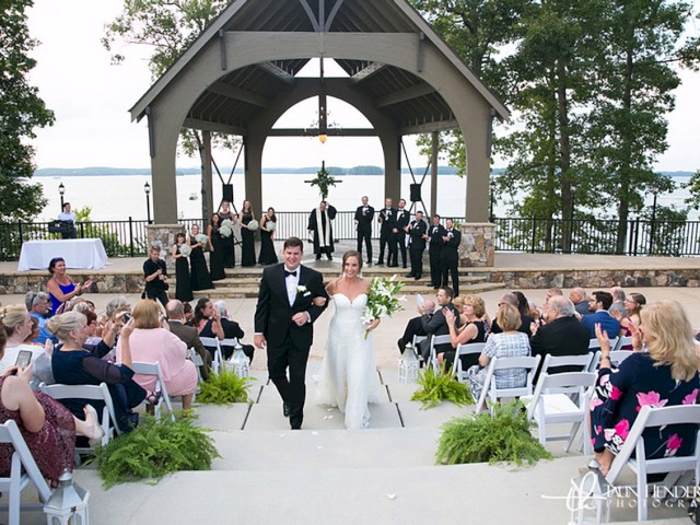 A bride and groom walk down the aisle outside, under a pavilion, with guests seated on either side, and bridesmaids and groomsmen in the background.