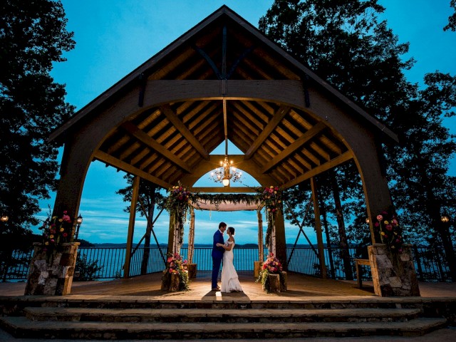 A couple is standing under a wooden pavilion with flowers, surrounded by trees at dusk. The scene has a romantic and serene ambiance.