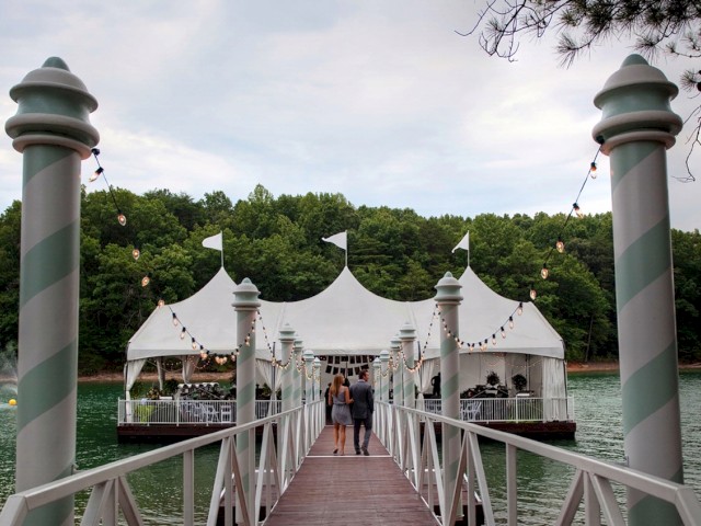 A couple walks along a dock towards a white tented pavilion on the water, surrounded by greenery, with string lights hanging above.