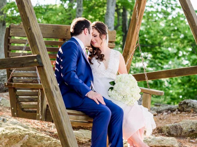 A couple in wedding attire sits on a wooden swing, surrounded by nature. The bride holds a bouquet of white flowers, and they share an intimate moment.