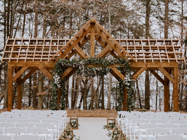 An outdoor wedding setup with white chairs in rows facing a decorative wooden arch adorned with greenery, set in a wooded area.