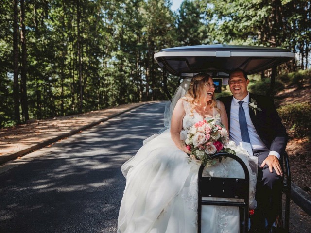 A bride and groom are sitting in a golf cart on a wooded road, smiling and holding a bouquet of flowers.
