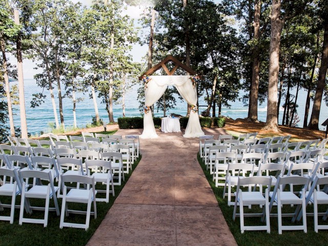 An outdoor wedding setup with white chairs arranged on either side of a walkway leading to an altar with draped fabric, surrounded by trees and a view of a lake.