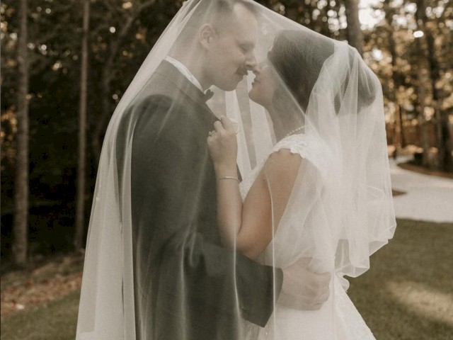 A couple under a veil embracing outdoors, with trees in the background, creating a romantic and serene atmosphere.