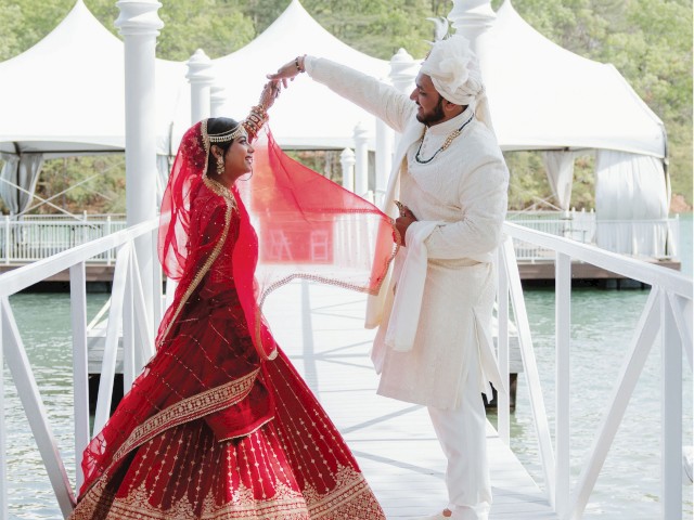 A couple in traditional attire dances on a pier with white tents and trees in the background.