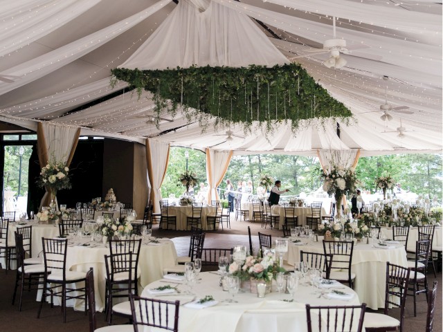 An elegant wedding reception tent with draped ceiling, hanging greenery, and tables set with floral arrangements and fine dinnerware.