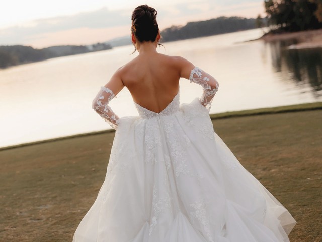 A person in a white wedding dress stands by a serene lake at sunset, with trees in the background, arms raised slightly.