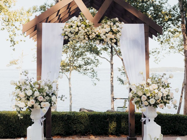 An outdoor wedding arch decorated with white flowers and drapery, set against a lakeside backdrop with trees in the distance.