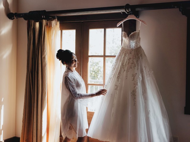 A woman stands by a window, holding a white wedding dress hanging from a rod, sunlight streaming in.