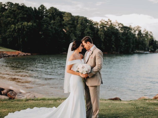A couple in wedding attire embraces by a scenic lake, surrounded by greenery and a serene sky.