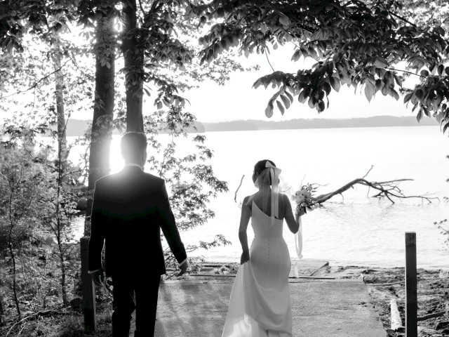 A couple walking towards a serene lake, surrounded by trees, in a black and white setting, possibly a wedding scene.